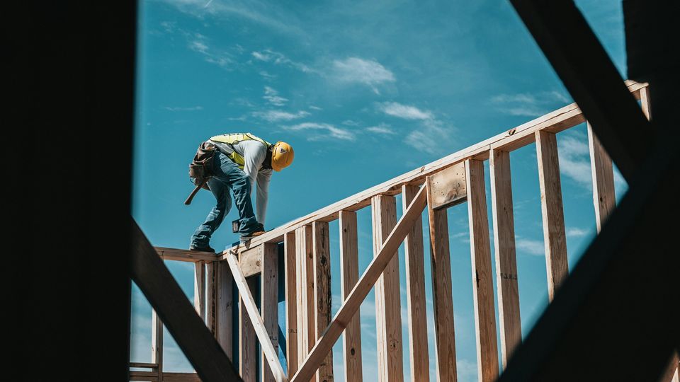 A construction working in a hi-vis vest and yellow hard hard crouches on top of a timber frame wall, securing its upper joints.
