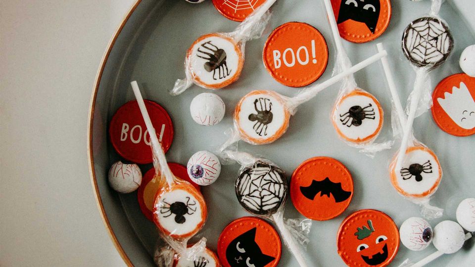 A grey bowl of Halloween candy, including foil covered eyeballs, lolly pops and orange coins with black bat figures featured.