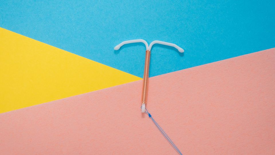 A T-shaped copper IUD, against a blue, yellow and pink triangular background.
