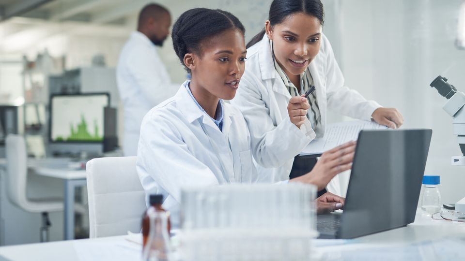 Two female scientists in white lab coats collaborating while looking at a laptop in a modern laboratory setting, surrounded by lab equipment and digital data displays, symbolizing the use of a software platform in scientific research.