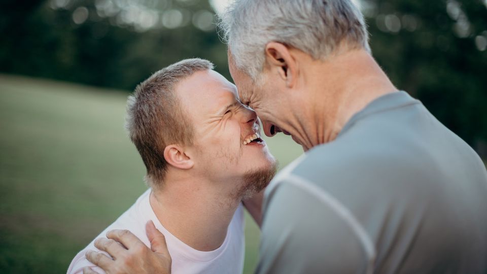 Young man with Down syndrome smiling and sharing a joyful moment with an older adult outdoors.