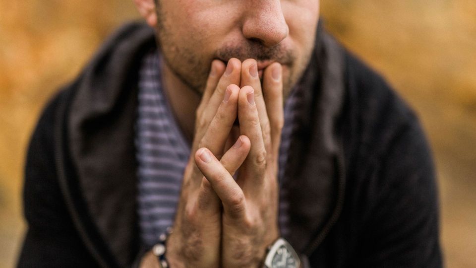 A man waits with his hands pressed together, resting against his face, looking thoughtful.