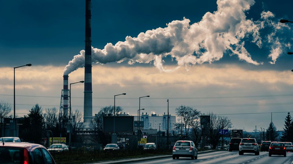 Cars on a highway driving past a factory emitting white smoke.