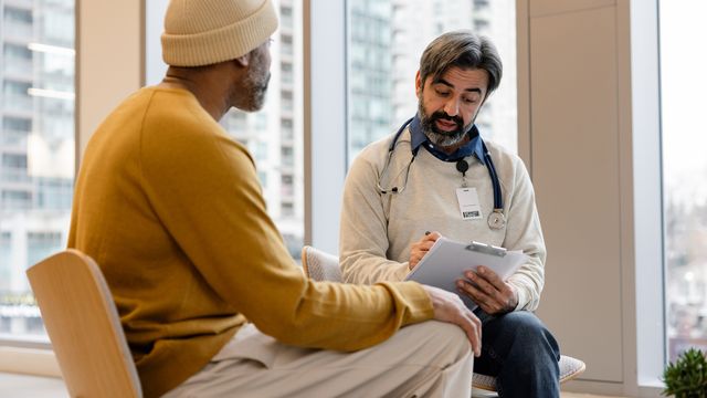 A seated male cancer patient speaking with a male doctor with a clipboard in front of a window with natural light. 