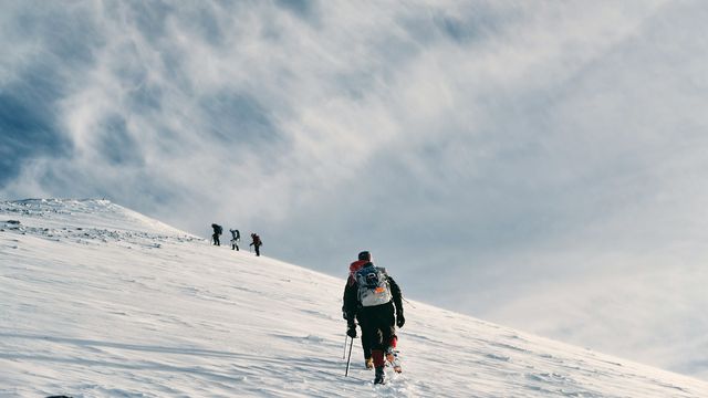 Climbers ascending a snow-covered mountain slope under a dramatic, cloud-filled sky. 