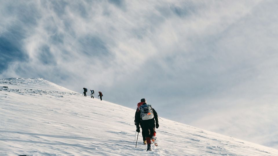 Climbers ascending a snow-covered mountain slope under a dramatic, cloud-filled sky.
