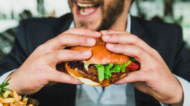 A smiling man in a suit, holding up a large burger and preparing to bite into it.  