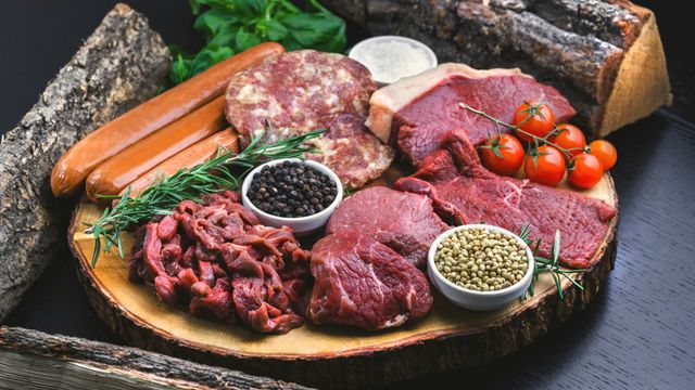 Different cuts of raw meat and sausages lying on a log-style plate, surrounded by herbs and spices. 