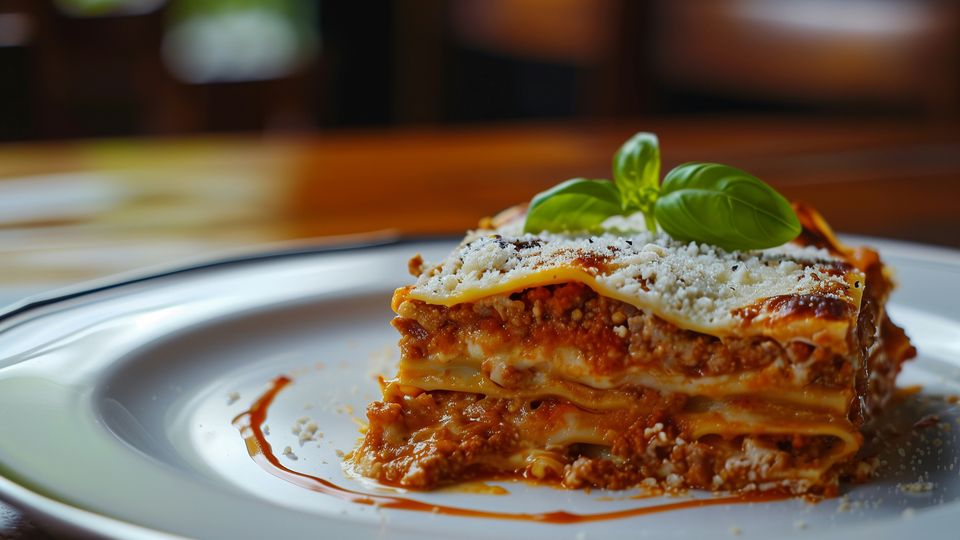 A portion of meat lasagna on a white plate, topped with a basil leaf.