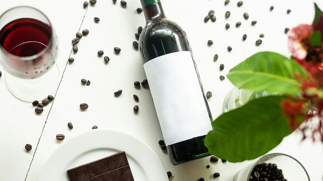 A glass of red wine, a wine bottle, a white plate with a piece of dark chocolate on it, a green plant and a small cup of coffee beans, all sat on a white wooden surface with some scattered coffee beans around them. 