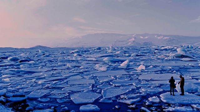 A landscape photograph of breaking ice sheets in Arctic waters. Two people stand on a large sheet in the bottom-right of the photograph. 