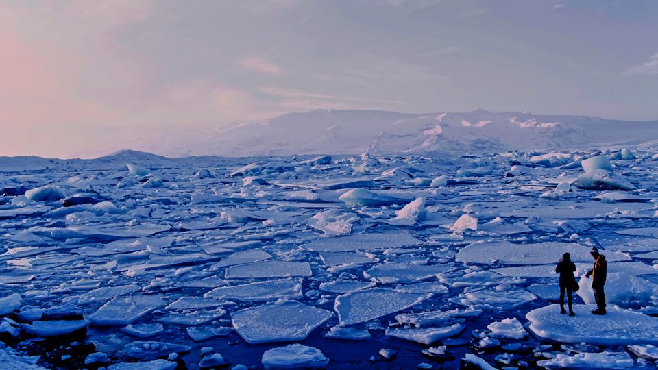 A landscape photograph of breaking ice sheets in Arctic waters. Two people stand on a large sheet in the bottom-right of the photograph.