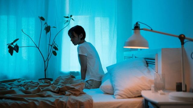 Woman sitting on bed in dim lighting, appearing fatigued, representing symptoms of long COVID. 