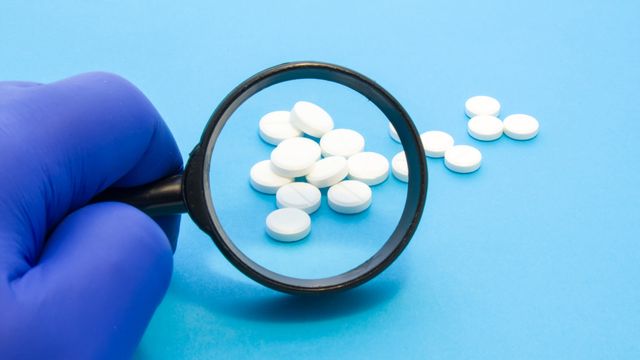 Gloved hand holding magnifying glass over white metformin tablets on a blue background. 