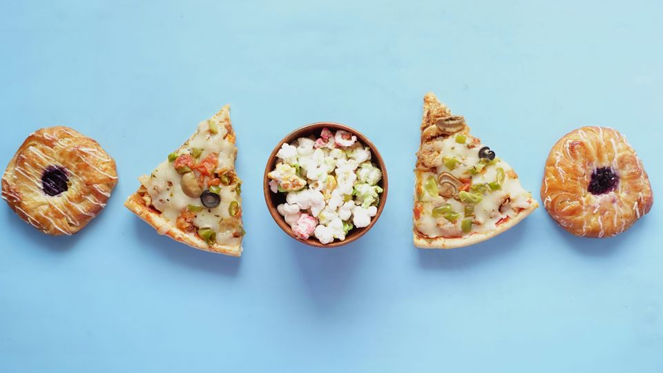 Two donuts, two slices of pizza and a bowl of popcorn, sitting on a blue background.