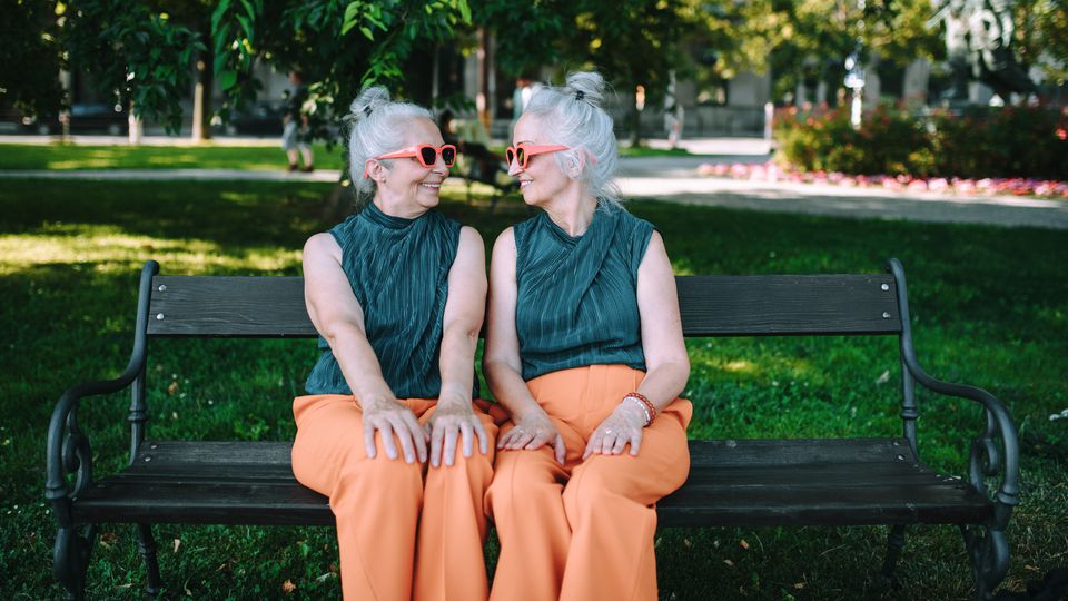 Identical twins in matching outfits smiling at each other on a park bench, symbolizing nature vs nurture.