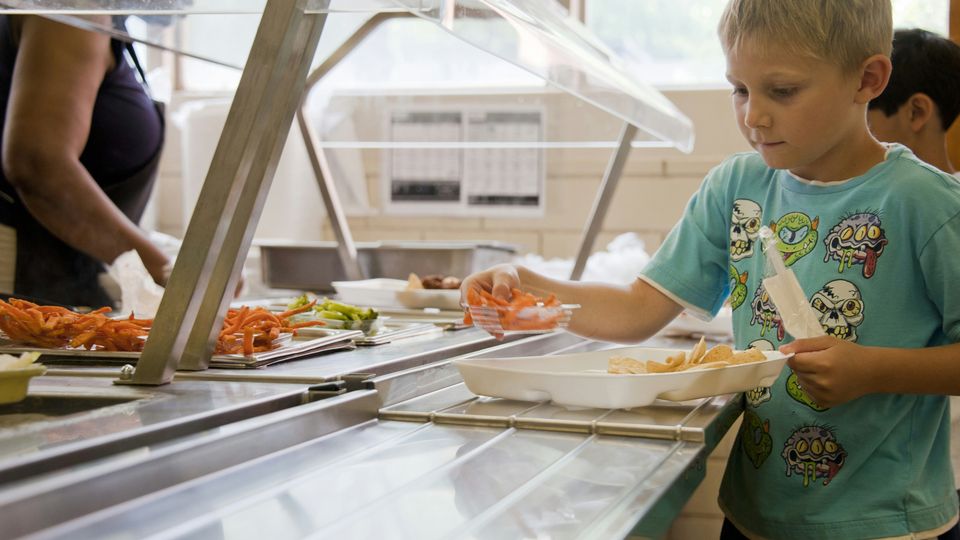 A boy in a blue cartoon top picking up items from a school cafeteria platter and placing them on his tray.
