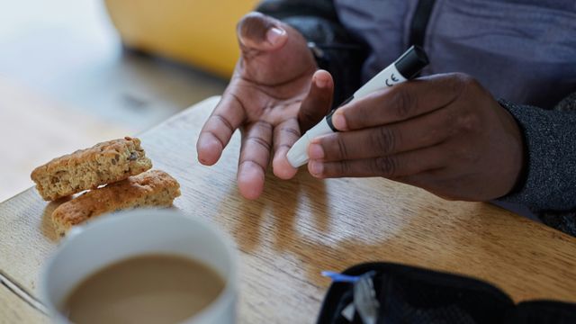 A man sat at a small table doing a fingerprick blood sugar test. In front of him is a cup of tea and two small cookies. 