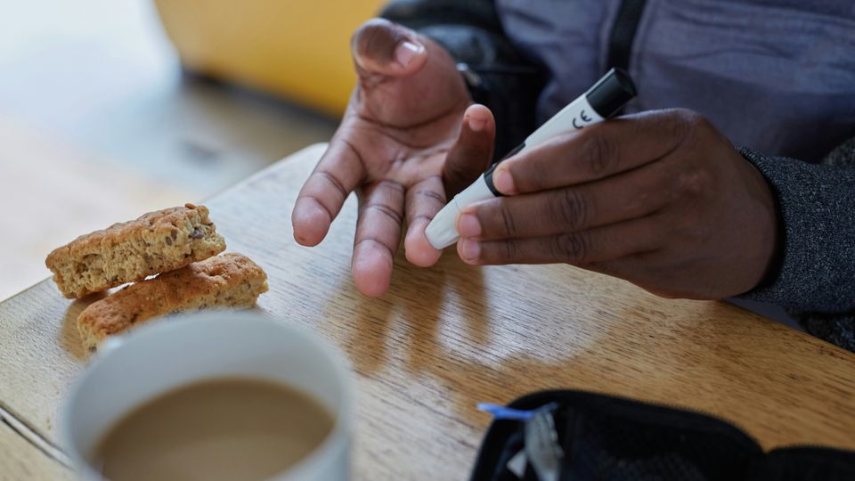 A man sat at a small table doing a fingerprick blood sugar test. In front of him is a cup of tea and two small cookies.