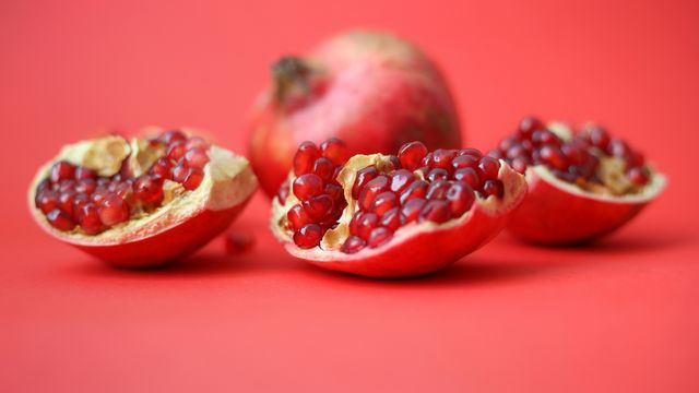 Three wedges of pomegranate sitting in front of a whole pomegranate, sitting against a light red background. 