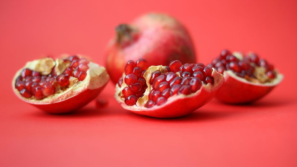 Three wedges of pomegranate sitting in front of a whole pomegranate, sitting against a light red background.