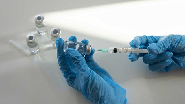 Close up of vaccine vial and syringe, with liquid being drawn up by person wearing blue gloves. 