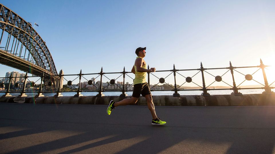 A man running alongside a river in a city, with a large bridge in the background.