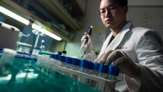 Male scientist in lab coat using a manual pipette, sitting with a rack of sample tubes. 