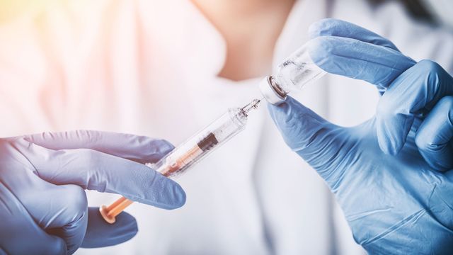 Close up of scientist wearing blue nitrile gloves, drawing liquid from a vial into a syringe. 