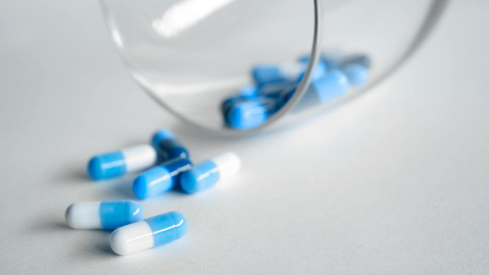Blue and white antibiotic capsules spilling from a glass container onto a white surface. 