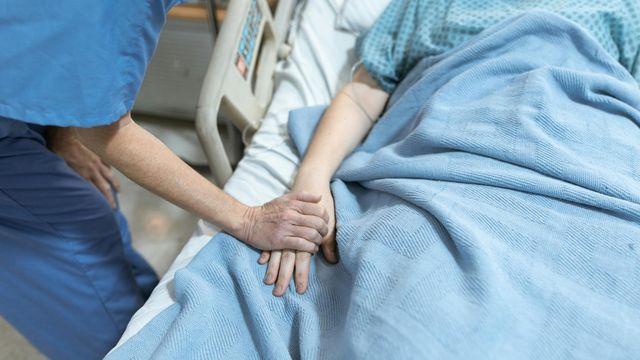 A hospital patient lies in bed covered with a blue blanket while a healthcare worker holds their hand 