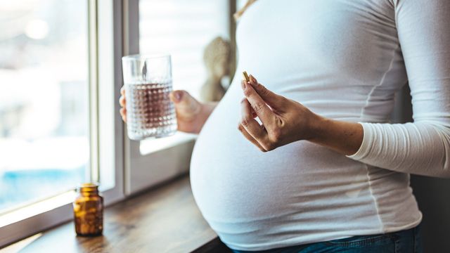 Pregnant woman holding a glass of water and prenatal supplement near a sunny window. 
