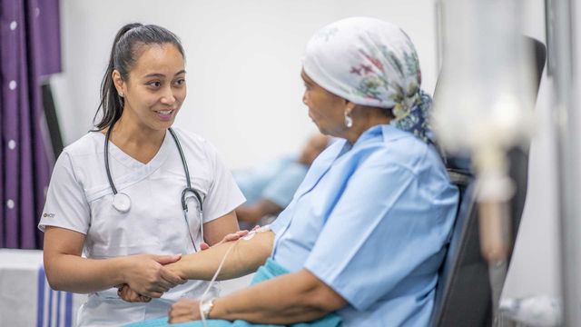 Female doctor speaking with a female cancer patient wearing a headscarf, who is raising her arm with an IV cannula inserted. 