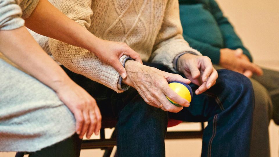 Close-up of an elderly person sitting and holding a small yellow and blue ball, with a caregiver offering support by holding their arm, illustrating care in a nursing home setting.