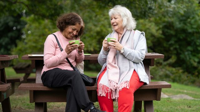 Two older women sitting on a park bench, smiling and laughing. They each hold a ceramic teacup in their hands. 