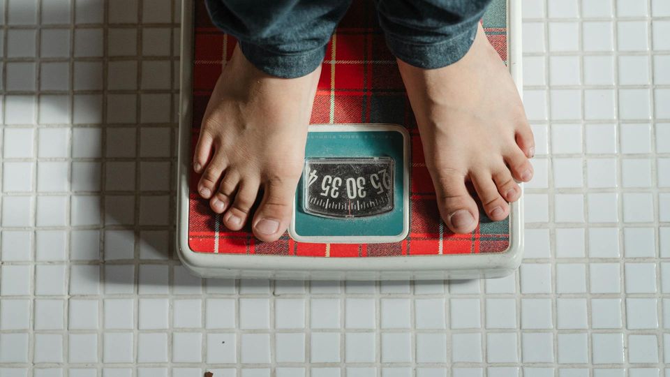 Person standing barefoot on a red bathroom scale, measuring body weight on a tiled floor, representing weight loss or weight management.