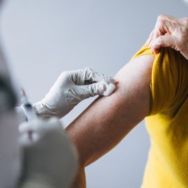 Healthcare professional wearing gloves preparing to administer a vaccine injection into the upper arm of an older adult wearing a yellow shirt. 