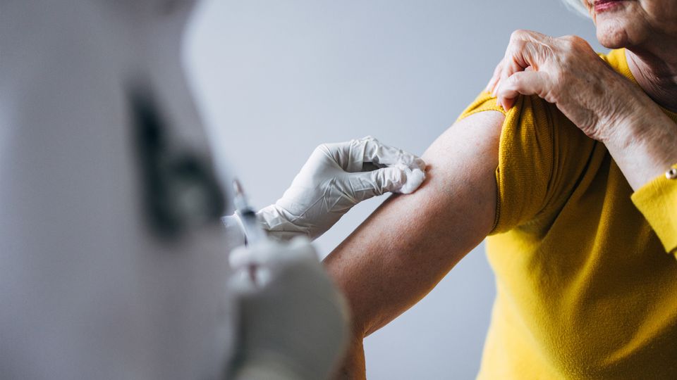 Healthcare professional wearing gloves preparing to administer a vaccine injection into the upper arm of an older adult wearing a yellow shirt.