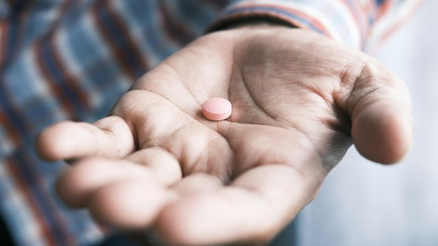 An older man holding a small pink pill in his outstretched hand. 