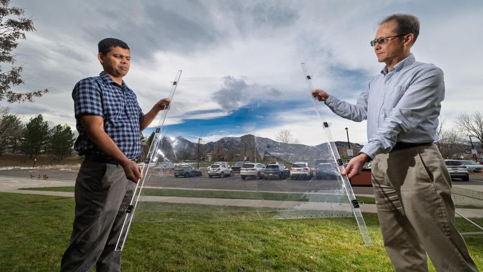 Two men holding a large transparent panel outside. Through the panel you can see a car park and some mountain peaks in the background.
