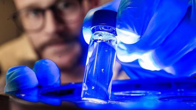 A researcher holds a small sample vial full of liquid over a bright blue LED, causing it to glow. 