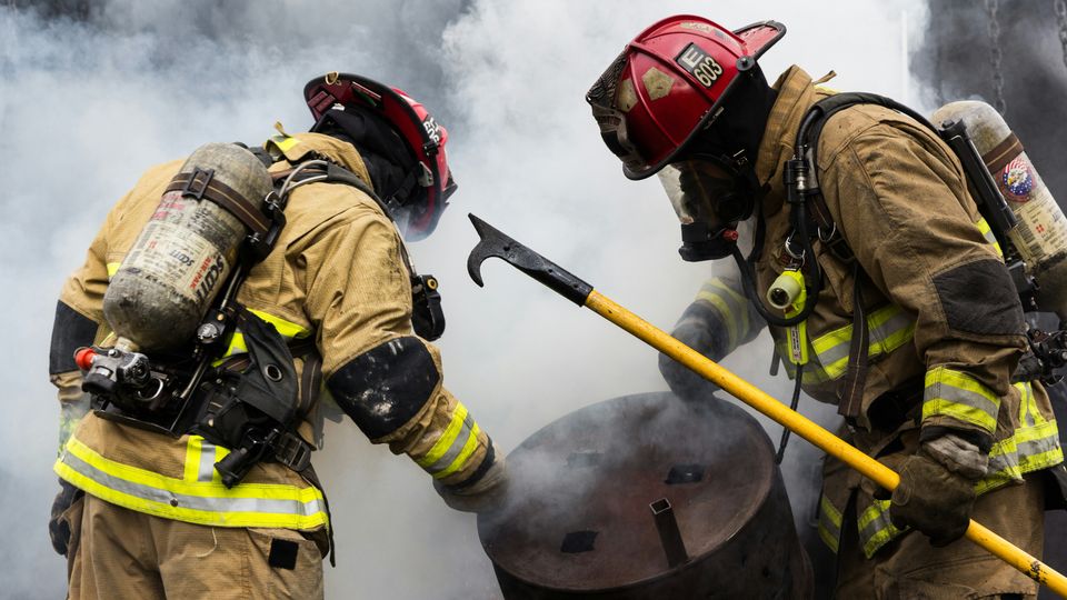 Two firefighters in protective gear carrying a large, rusty, metal cylinder. The background of the image is covered in white smoke.