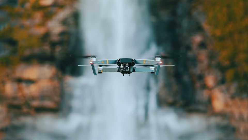 A silver drone hovering in front of a waterfall and stream.