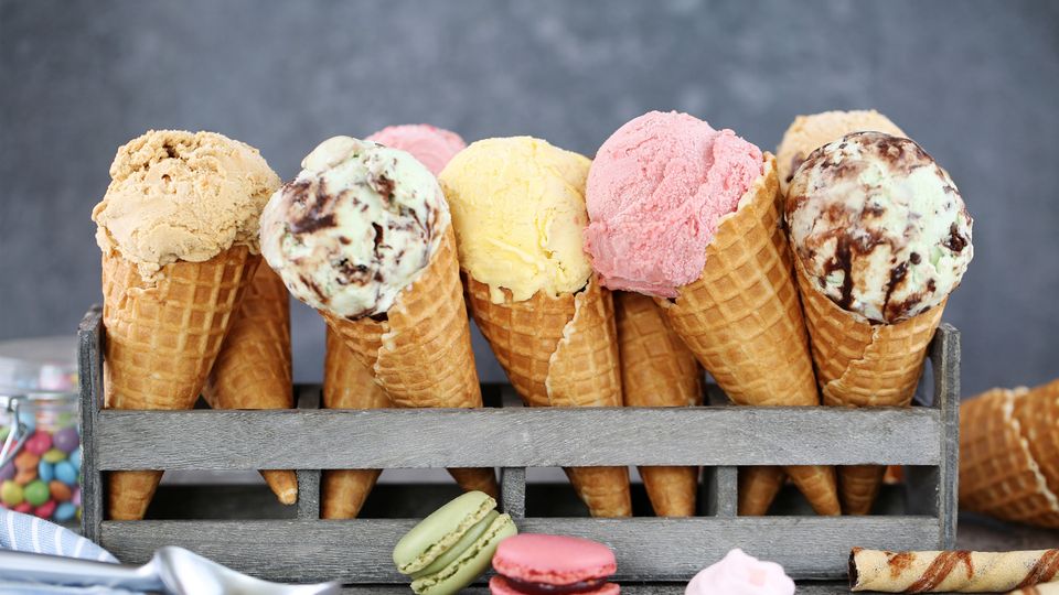Assorted scoops of ice cream in waffle cones displayed in a holder, with macarons and sweets in the foreground, illustrating a concept relevant to rheology in food texture and flow behavior.