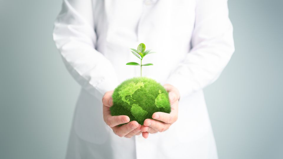 A researcher in a white lab coat holding a fuzzy green ball, edited to look like the Earth, with a small sapling growing out the top of the Earth.
