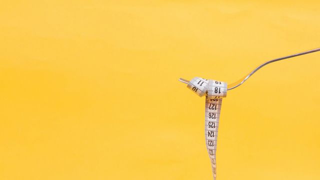 Measuring tape twirled on a fork against yellow background, symbolizing obesity and dieting. 