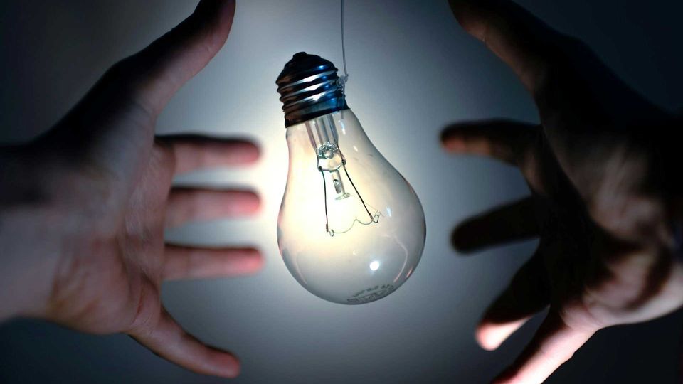 Hands surrounding a glowing light bulb suspended against a dark background.