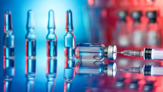 Vaccine vials on a mirrored surface with a blue and red haze, with a needle inserted into an overturned vial. 