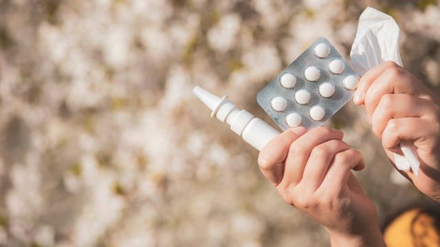 Hands of a person holding nasal spray, tablets, and tissues against a blurred beige background. 