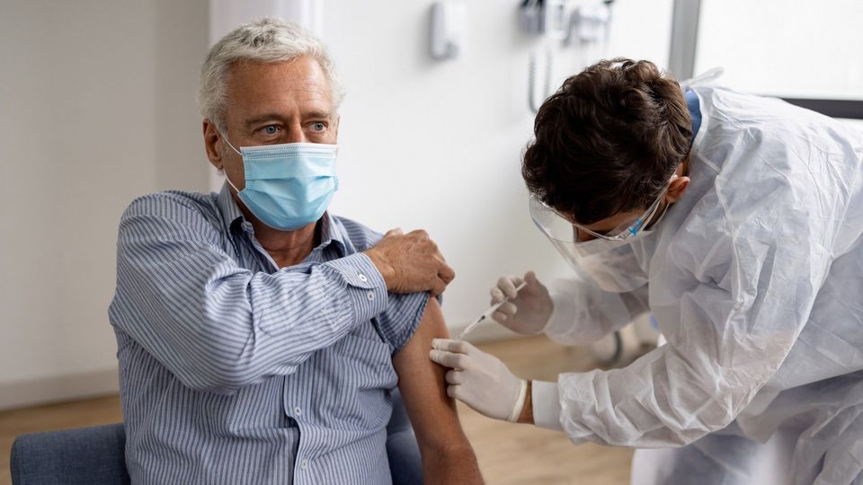 Man sat in a clinic room wearing a face mask, with his left sleeve rolled up, receiving a vaccine shot from a clinician.